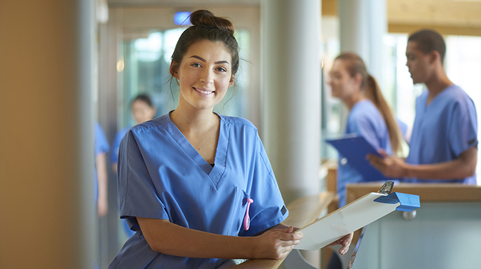 A medical assistant stands with a clipboard at a nurses station